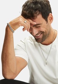 Silberne Kettenarmband, Silberne Anhänger-Halskette, weißes strukturiertes T-Shirt. Männliche Figur mit dunklen, lockigen Haaren und heller Haut, die die Hand an die Stirn legt.