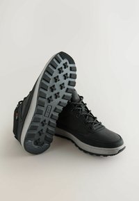 Mustat vaelluskengät, joissa on teksturoitu päällinen, harmaa välikerros ja kestävä kumipohja, jossa on syvä urakuvio. Nauhat ovat heijastavat.