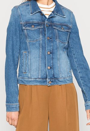 Blaue Jeansjacke mit Knopfverschluss, getragen über einem gestreiften weißen und beigefarbenen Hemd und brauner Hose mit hoher Taille.
