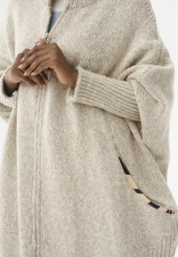 Beżowy sweter z dzianiny z zamkiem z przodu, oversize'owe rękawy, prążkowane mankiety i ozdobna kieszeń w paski. Miękka, teksturowana tkanina.