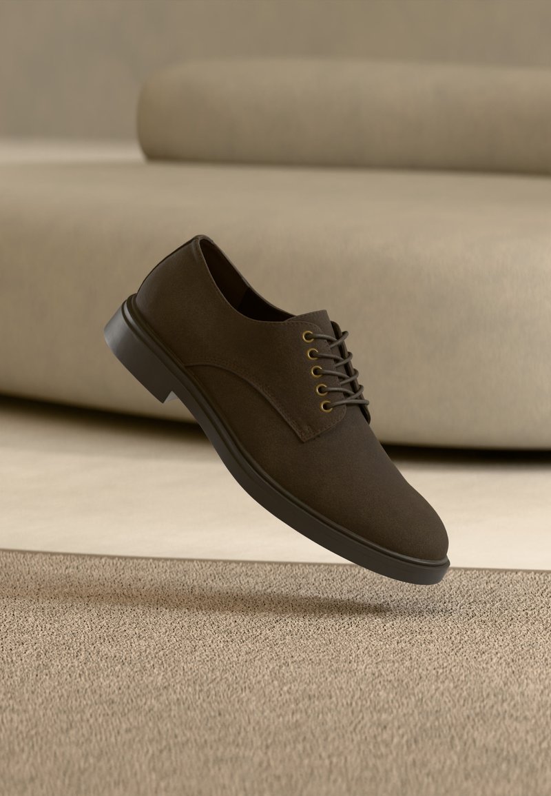 Καφέ σκούρο δερμάτινο ανδρικό παπούτσι με κορδόνια αιωρείται πάνω από μπεζ χαλί, με μπεζ καναπέ θολωμένο στο φόντο.