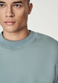 Sudadera azul claro con cuello redondo acanalado y textura suave, que presenta un corte relajado y detalles de diseño mínimos.