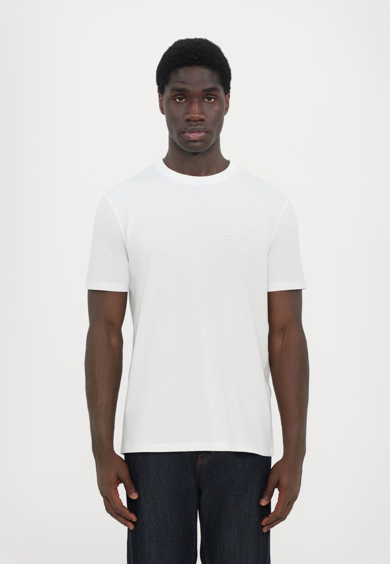 Hvit t-skjorte med rund hals laget av bomull, med korte ermer og en subtil logo på brystet. Kombinert med mørkeblå denimjeans.
