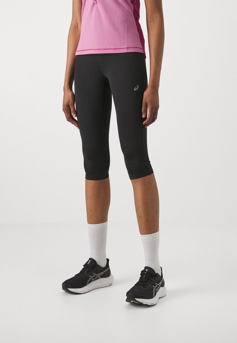 Leggings sportivi neri con un design aderente, lunghezza cropped e tessuto elasticizzato. Indossati con una maglietta rosa, sneakers nere e calzini bianchi.