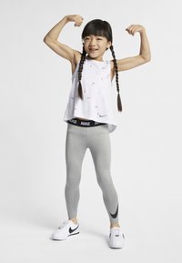 Άσπρο αμάνικο τοπ με πολύχρωμα λογότυπα Nike, γκρι κολάν και άσπρα αθλητικά παπούτσια με μαύρες λεπτομέρειες. Παιδί που λυγίζει τα χέρια του με αυτοπεποίθηση.