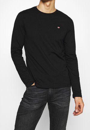 Homme portant un t-shirt noir à manches longues avec un petit logo de drapeau rouge et blanc sur la poitrine et un jean gris foncé.