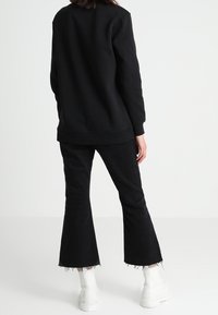 Osoba stojící zády, oblečená v černých rozšířených kalhotách s neupravenými lemy, černém dlouhém rukávu a bílých kotníkových botách.