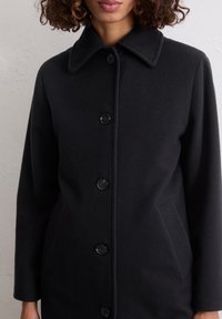 Frau trägt einen schwarzen, knopfgeknöpften Wollmantel mit spitzem Kragen und seitlichen Taschen vor einem schlichten, hellen Hintergrund.