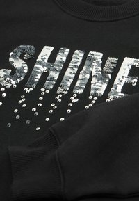 Czarna bluza z kapturem z napisem "SHINE" w dużych srebrnych cekinach, z dodatkowymi małymi srebrnymi cekinami spadającymi poniżej. Prążkowane mankiety i dekolt.