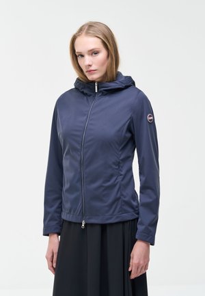 Jonge vrouw die een marineblauwe capuchonvest met rits draagt, met een klein rond logo op de linker mouw, staande tegen een effen witte achtergrond.