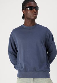 Navy blue sweatshirt in soft fabric with long sleeves and a round neckline, featuring a ribbed hem and cuffs. Worn with black sunglasses.