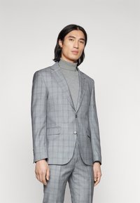 Completo grigio a quadri con vestibilità su misura, caratterizzato da revers a lancia, chiusura a un bottone e pantaloni coordinati, indossato sopra un dolcevita grigio.