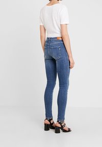 Jeans azul ajustados con cintura alta, hechos de mezclilla, con bolsillos traseros y un parche de cuero marrón en la cintura. Llevados con sandalias de tacón negras.