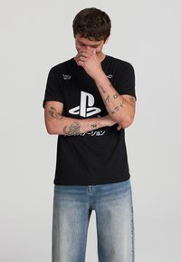 T-shirt in cotone nero con un grande logo PlayStation bianco e testo in giapponese. Vestibilità standard con maniche corte, design minimalista.