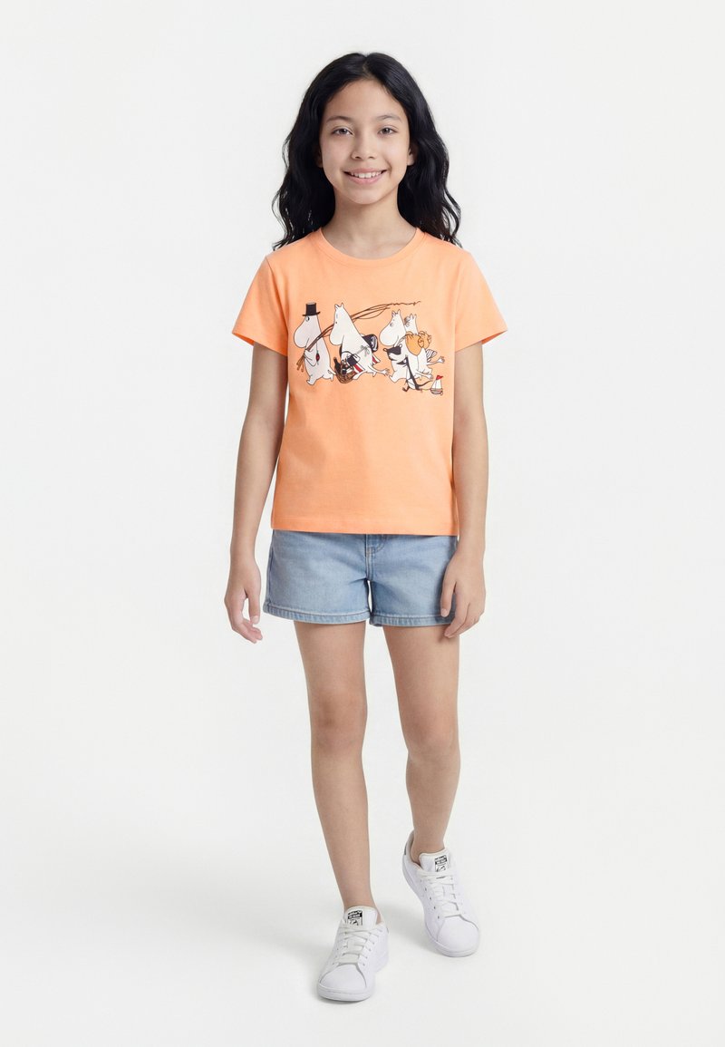 Κορίτσι που χαμογελά, στέκεται, φοράει πορτοκαλί μπλούζα με τυπωμένο καρτούν, τζιν σορτς και λευκά αθλητικά παπούτσια σε λευκό φόντο.