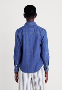 Sinine teksashirt pikkade varrukatega, nööbitud mansettidega ja terava kraega. Omab kaardus serva ja nähtavaid õmblusdetails.