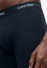 Fekete férfi boxeralsók sima anyagból, kontrasztos derekúval, amelyen fehér "Calvin Klein" logó található. Közeli nézet, amely hangsúlyozza a szabást.