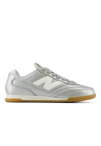Silver athletic shoes with a smooth texture, featuring a white “N” logo, gray laces, and a gum rubber sole. Streamlined design with subtle stitching.