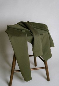 Felpa verde oliva appoggiata su uno sgabello di legno contro un semplice muro grigio, illuminata da una luce naturale soffusa.