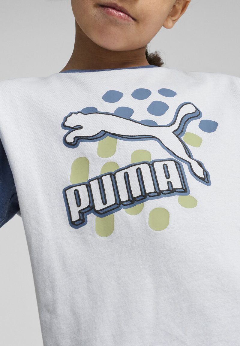 Balta kokvilnas T-kreklā ar zilām piedurknēm, uz kura ir Puma logo bieza burtiem un lecoša kaķa grafika, uz zaļiem un zilajiem punktiem.