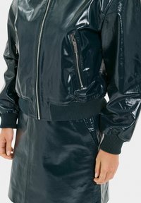 Bomber jacket verde scuro e lucido con polsini e orlo a coste, dotato di tasca sul petto con zip e silhouette snella.