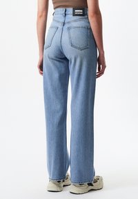 Personne portant un jean droit taille haute bleu clair avec poches arrière, associé à des baskets beiges et un haut marron sans manches.