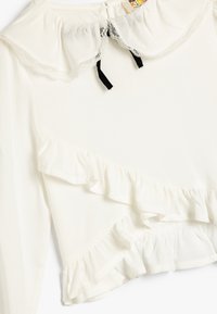Weiße Bluse mit einem gerüschten Ausschnitt mit Spitzenbesatz, vertikalen Rüschendetails an der Vorderseite und schwarzen Satindetails.