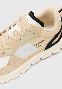 Chaussure de sport beige fabriquée en mesh et en daim, avec des accents blancs et noirs, des détails texturés et une semelle rembourrée.