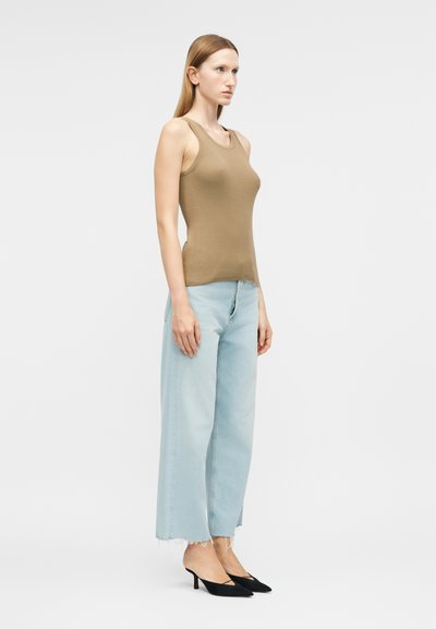 Γυναίκα που στέκεται προφίλ φορώντας ένα καφέ αμάνικο ριμπ μπλουζάκι, ανοιχτό μπλε τζιν παντελόνι με φαρδιά πόδια και μαύρες μυτερές γόβες στιλέτο.