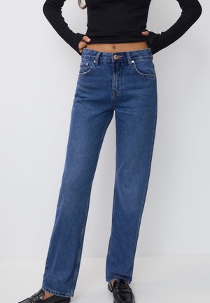 Woman wearing high-waisted blue straight-leg jeans, black cropped sweater, and black loafers, standing with hands on hips against white wall.