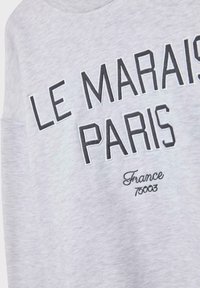 Ljust grått tyg med svart broderad text som läser "LE MARAIS PARIS" och med mindre kursiv text "Frankrike 75003" nedanför.