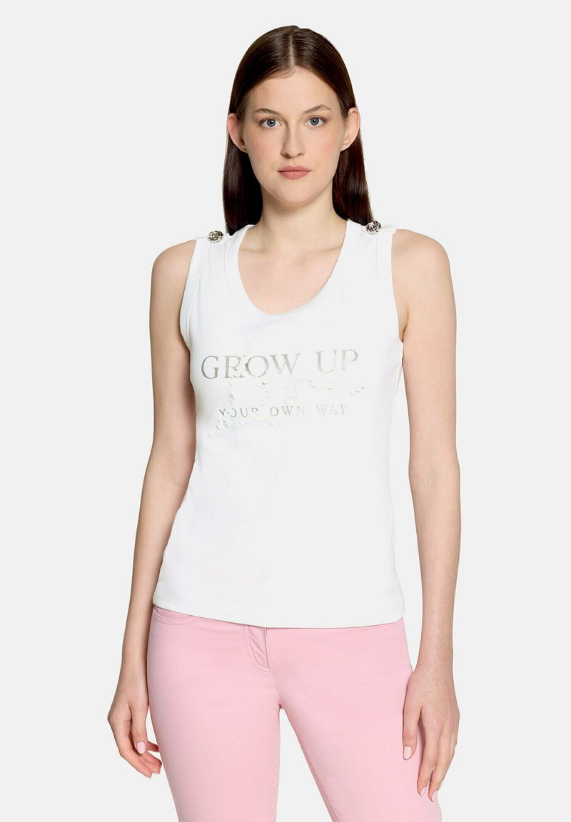 Femme aux cheveux bruns et raides porte un haut blanc sans manches avec des boutons décoratifs et un texte argenté, assorti à un pantalon rose clair.