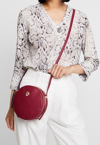 Borsa a tracolla rotonda in pelle bordeaux con hardware dorato e chiusura con zip. Indossata con una maglietta a fantasia e pantaloni bianchi.