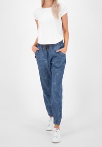 Blaue gemusterte Jogginghosen mit Kordelzugbund, ausgestattet mit Taschen und elastischen Bündchen, kombiniert mit einem schlichten weißen T-Shirt und weißen Sneakers.