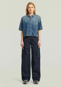 Egy nő áll, rövid ujjú, cropped denim ingben, széles szárú fekete cargo nadrágban és szürke sportcipőben, sima háttér előtt.