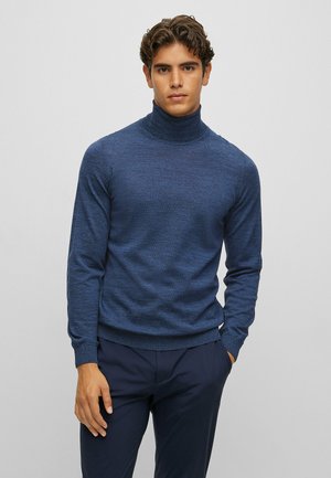 Pull à col roulé bleu marine en tissu texturé, avec des manches longues et une coupe ajustée. Associé à un pantalon sombre.