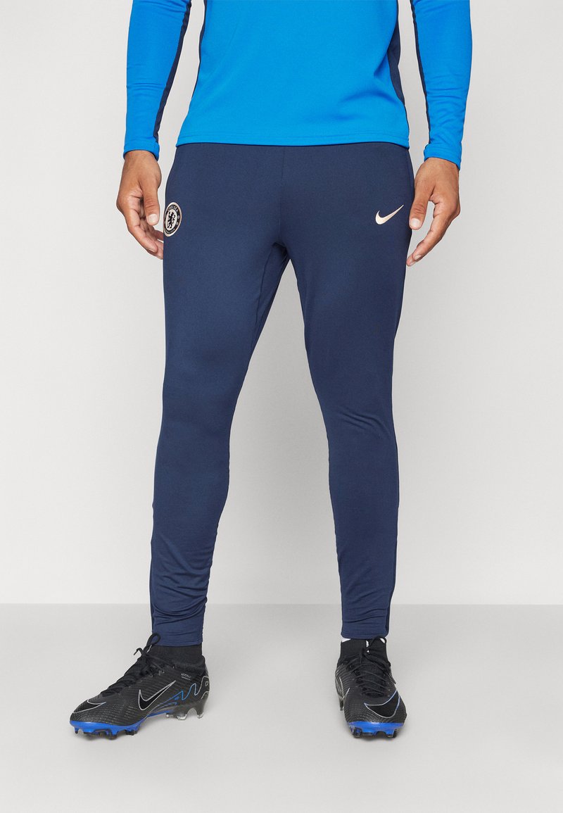 Granatowe spodnie sportowe o dopasowanym kroju, z logo Chelsea FC oraz białym znakiem Nike. W zestawie z czarnymi butami piłkarskimi.