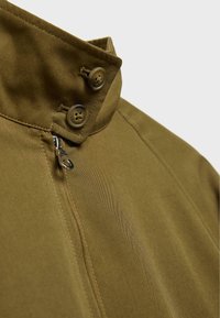 Khakifarbenes Jacket mit hohem Kragen, zwei Knöpfen, einem Reißverschluss und glatter Stoffstruktur mit sichtbaren Nähdetails.