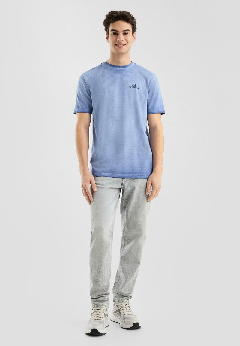 Lichtblauw t-shirt met korte mouwen en een ronde hals, voorzien van een klein geborduurd logo. Draagt met lichtgrijze jeans en grijze sneakers.