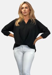 Czarna bluzka z długim rękawem, dekoltem w serek i węzłem z przodu, w połączeniu z białymi dopasowanymi spodniami. Gładka tekstura, minimalistyczny styl.