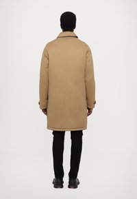 Long, padded coat in light brown fabric with a buttoned cuff detail. Worn over black pants and shoes, featuring a back collar design.