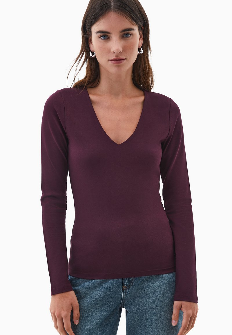 Giovane donna che indossa orecchini a cerchio d'argento, un top fitted a V color prugna con maniche lunghe e jeans blu, in piedi di fronte a uno sfondo bianco.