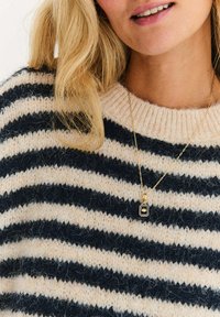 Granatowo-kremowy sweter w paski o miękkiej fakturze. Naszyjnik z złotym wisiorkiem w kształcie prostokąta.