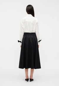 Λευκό πουκάμισο με μακριά μανίκια και πιέτες στο πίσω μέρος, σε συνδυασμό με μαύρη φούστα μήκους έως το γόνατο σε σχήμα Α, με φιόγκους στα μανίκια.