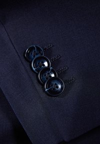 Tessuto di abito blu navy con quattro bottoni scuri, caratterizzato da una superficie liscia e fori a forma di fiore, cuciti con filo coordinato sul polsino.