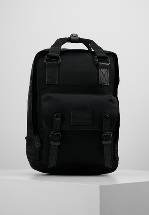 Czarny plecak z górnymi uchwytami, przednią kieszenią zabezpieczoną dwoma paskami z klamrami i prostokątną łatką na białym postumencie na szarym tle.