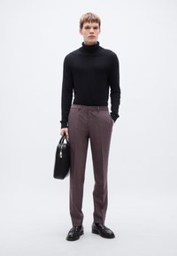 Μαύρο πουλόβερ με λαιμόκοψη ζιβάγκο, ραμμένο ανοιχτό μοβ παντελόνι και γυαλισμένα μαύρα μοκασίνια. Κρατάει μια μαύρη, δομημένη τσάντα χειρός. Απλό φόντο.