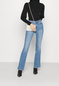 Zwarte geribbelde coltrui, lichtblauwe vloeiende denim jeans, witte geprofileerde handtas en zwarte enkellaarsjes, weergegeven tegen een neutrale achtergrond.