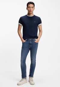 Tmavomodré tričko s okrúhlym výstrihom s detailmi svetlosivých manžiet, doplnené priliehavými modrými džínsami a béžovými teniskami, postávajúce s rukami v vreckách.