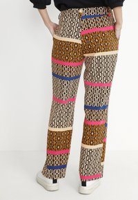 Kategorické kalhoty s multikolorovými vzory zahrnujícími odvážné tvary v černé, hnědé, růžové a modré barvě. Rovný střih s viditelnými detaily kapes.